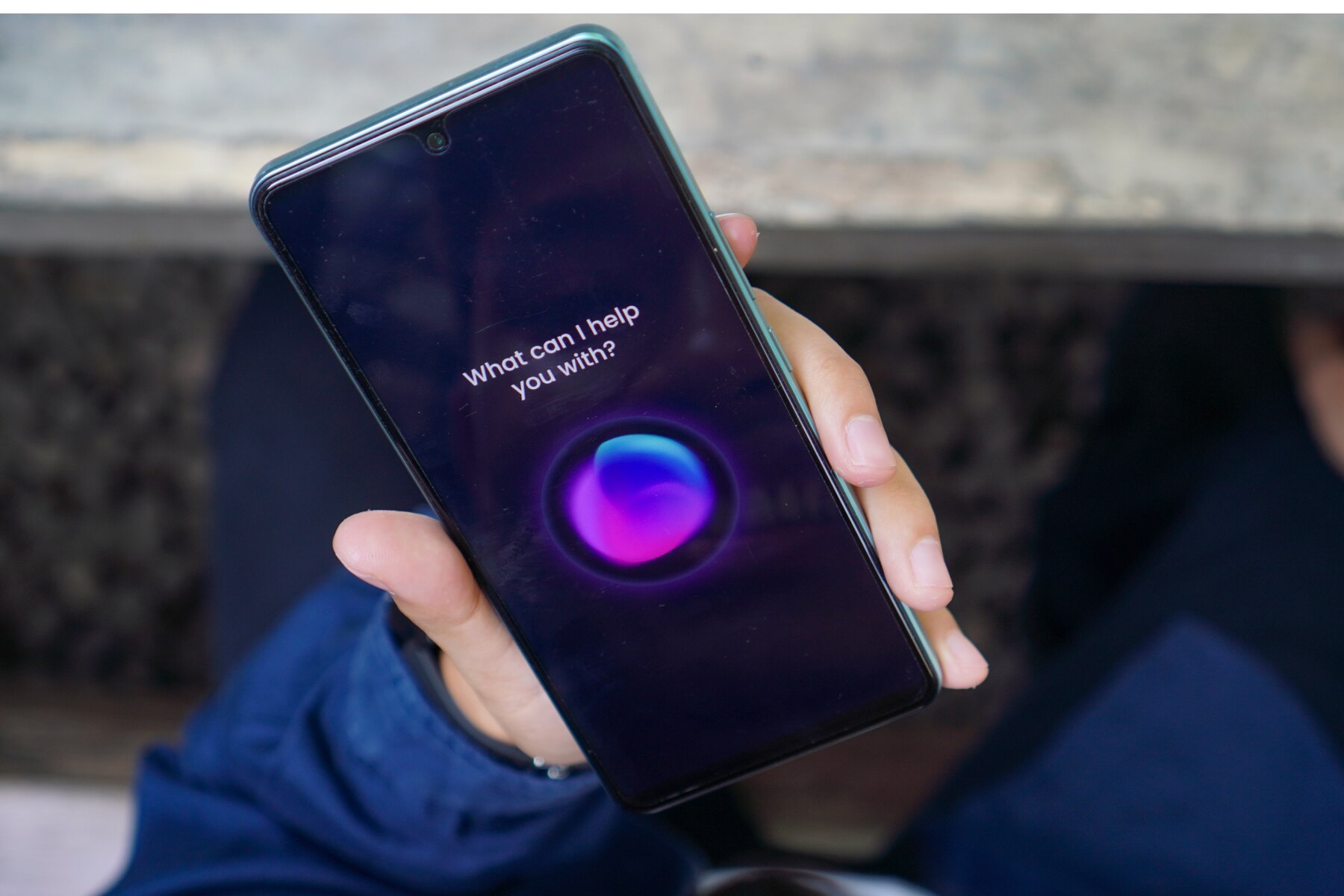 A person holding a smartphone displaying a virtual assistant interface with the text “What can I help you with"?