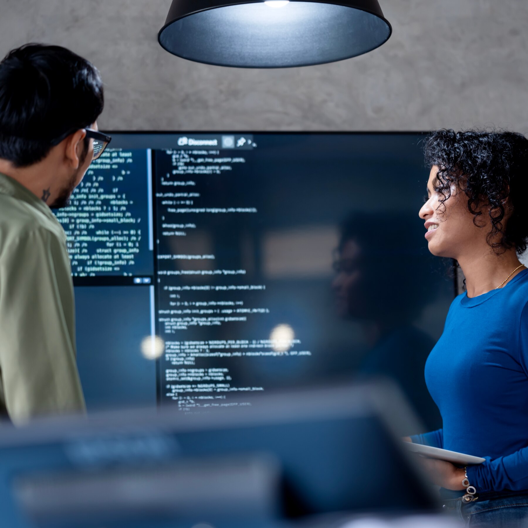 Two professionals discuss programming code displayed on a large screen, suggesting collaborative software development or code review in a modern workspace.