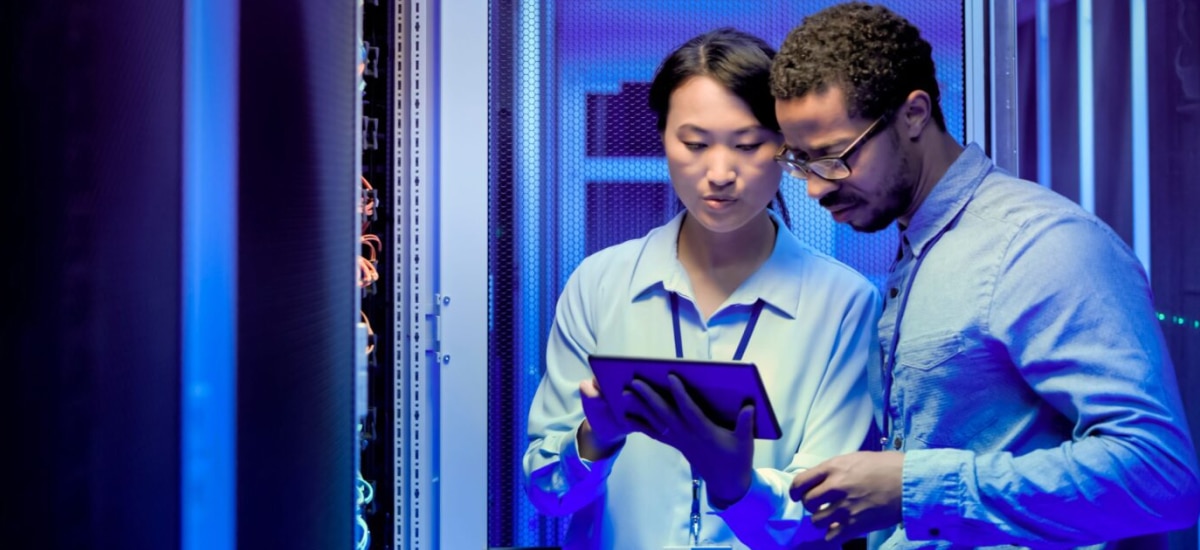 Two engineers reviewing data on a tablet inside a server room with blue lighting.